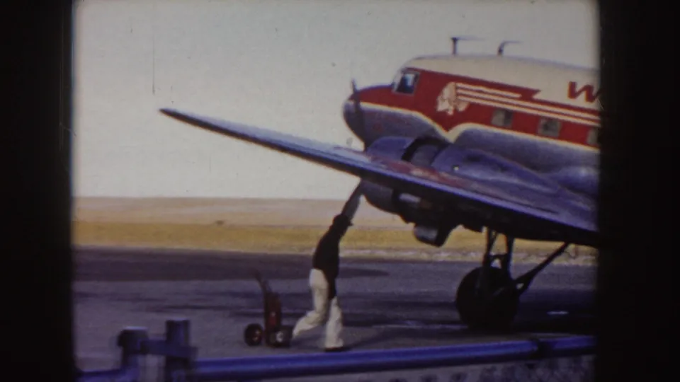 1955 Idaho 8mm frame showing the front section of a Western Airlines propeller plane on the tarmac, with a ground crew member and small cart near the wing.