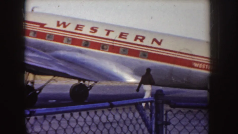 1955 Idaho 8mm frame showing the midsection of a Western Airlines aircraft, polished wing surface, and a person walking along the tarmac behind a chain-link fence.