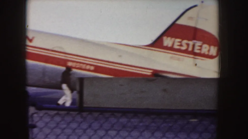 1955 Idaho frame showing the tail section and rear fuselage of a Western Airlines aircraft, with a ground crew member walking in front of the plane on the tarmac.