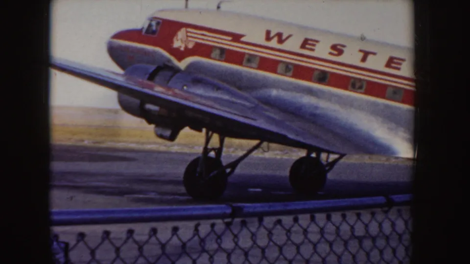 1955 Idaho home-movie frame showing the nose and wing of a Western Airlines DC-3 taxiing behind an airport fence.