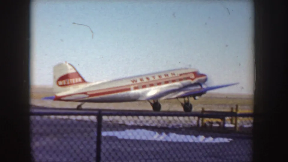 1955 Idaho frame with a Western Airlines DC-3 in side view on a snowy airport runway behind fencing.