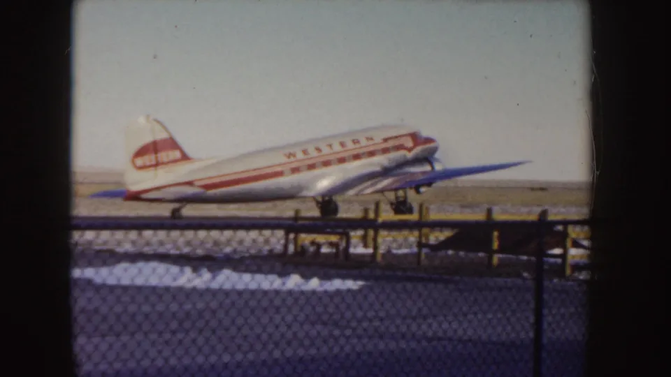 1955 Idaho home-movie view of a Western Airlines DC-3 moving along a snowy runway behind airport fencing.