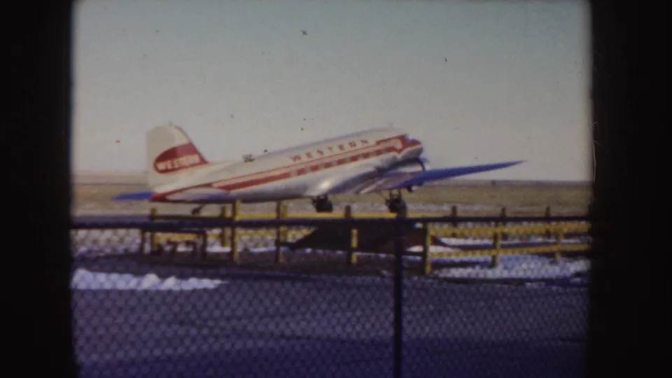 1955 Idaho frame showing a Western Airlines DC-3 taxiing behind a fence, with visible 8mm frame edge and faded color.