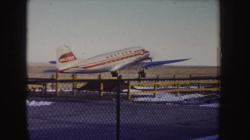 1955 Idaho 8mm frame showing a Western Airlines propeller plane near touchdown or taxiing on a snow-edged runway behind a fence.