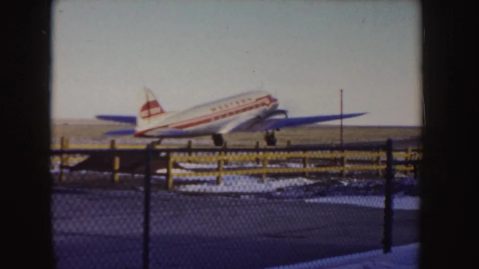 The Western Airlines propeller aircraft moves across a flat snowy Idaho airfield, filmed from behind a dark fence line.