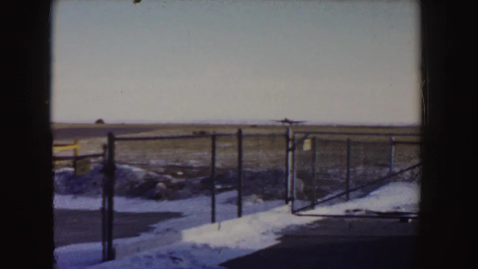 A distant propeller plane moves away across the open snowy airfield, with chain-link fencing and film borders framing the scene.