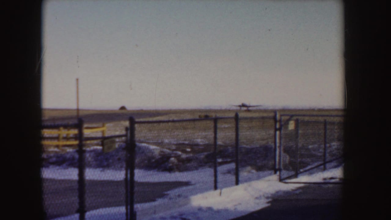 1955 Idaho 8mm frame showing a small aircraft near a snowy runway, viewed past a chain-link fence and yellow barrier.