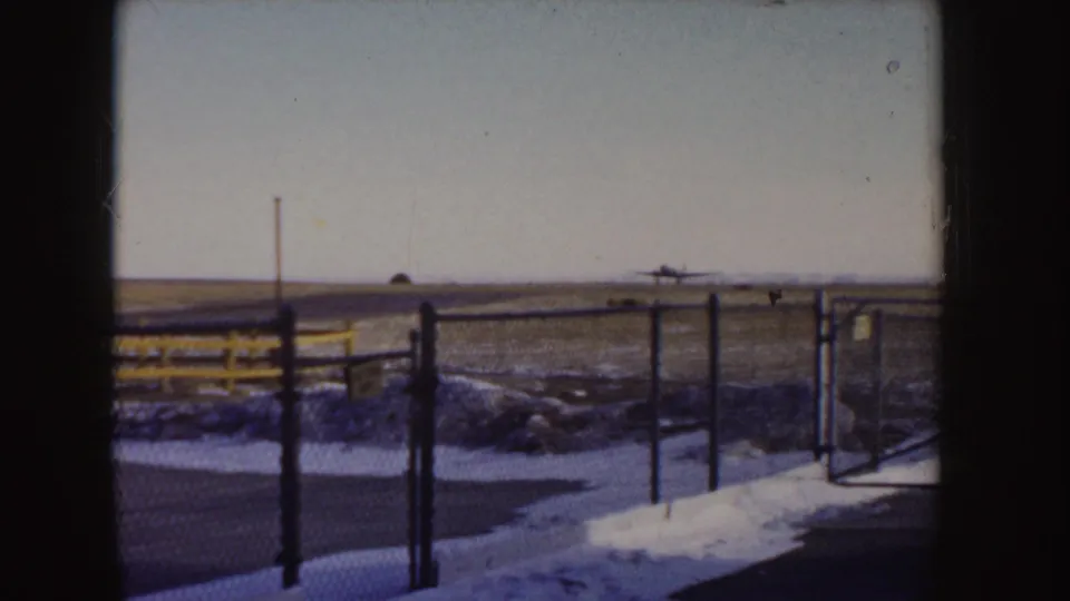 1955 Idaho 8mm frame with a small aircraft descending over a winter runway beyond fence posts and snow patches.