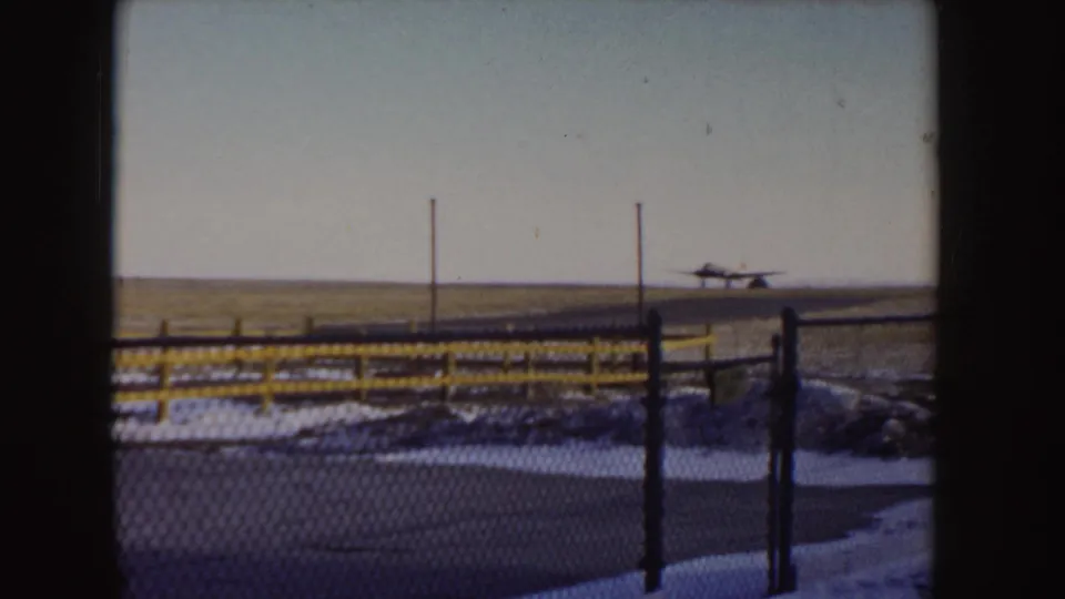 1955 Idaho 8mm frame showing the aircraft close to the snowy runway, seen past a yellow rail, fence, and utility poles.