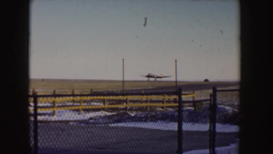 1955 Idaho 8mm frame of a small aircraft landing or taxiing along a snowy runway behind a chain-link fence and yellow rail.