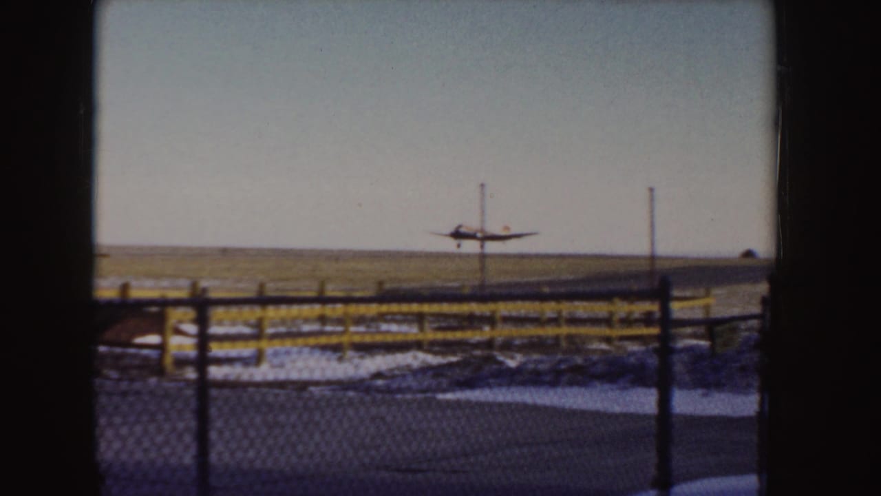 Small propeller plane lifting off beyond a chain-link fence and yellow rail.