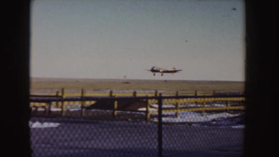 Small propeller plane continues its low climb over an open Idaho airfield, with fence rails and prairie ground below.