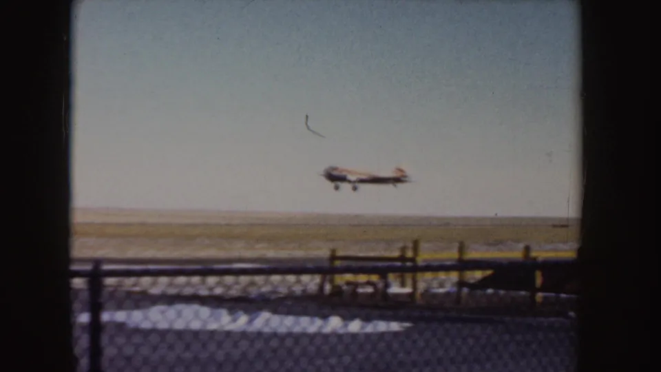 1950s 8mm view of a small propeller plane airborne over a flat Idaho airstrip, seen through foreground fencing.