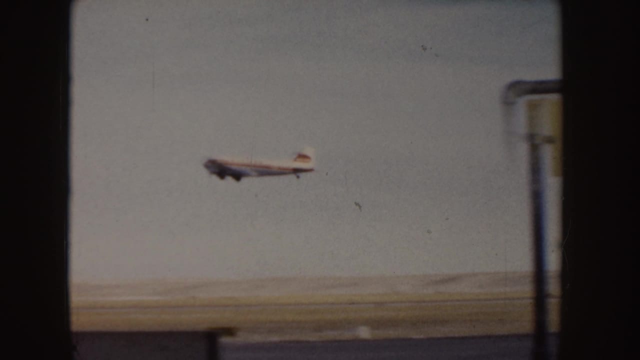 Small grey passenger plane climbing above a runway, seen through dark window framing.