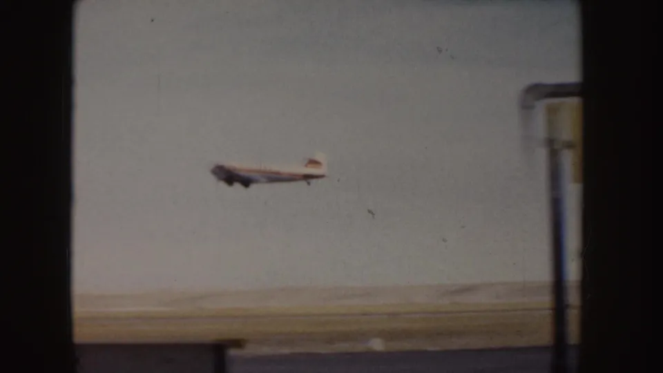 1955 Idaho 8mm frame of a small grey aircraft taking off beyond a runway, viewed through a shadowed airport-side opening.