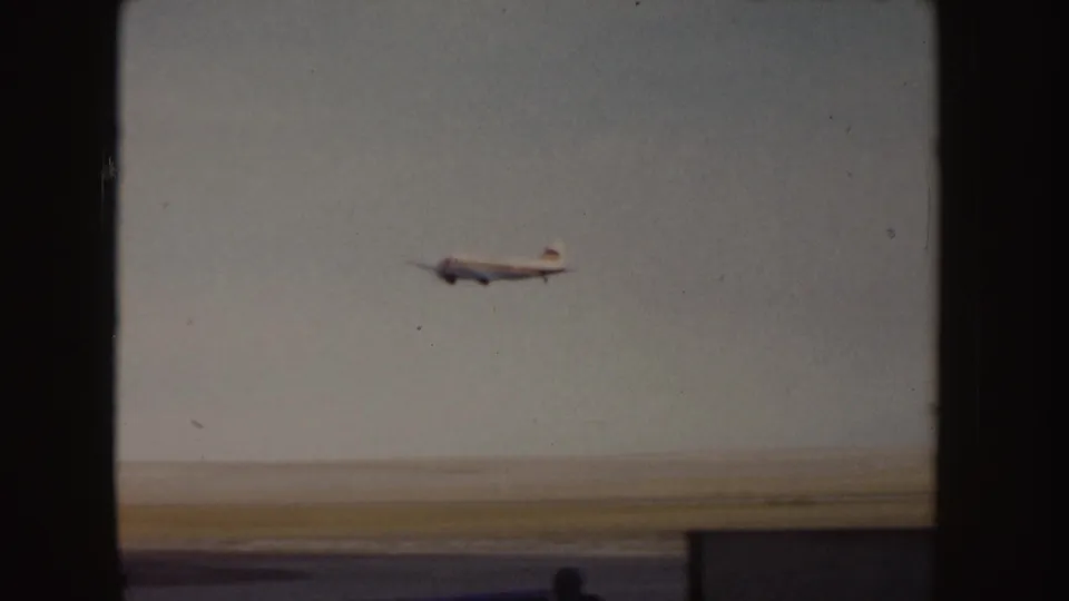 Small passenger plane climbs over the runway in 1955 Idaho footage, partly framed by a dark post and shadowed foreground.