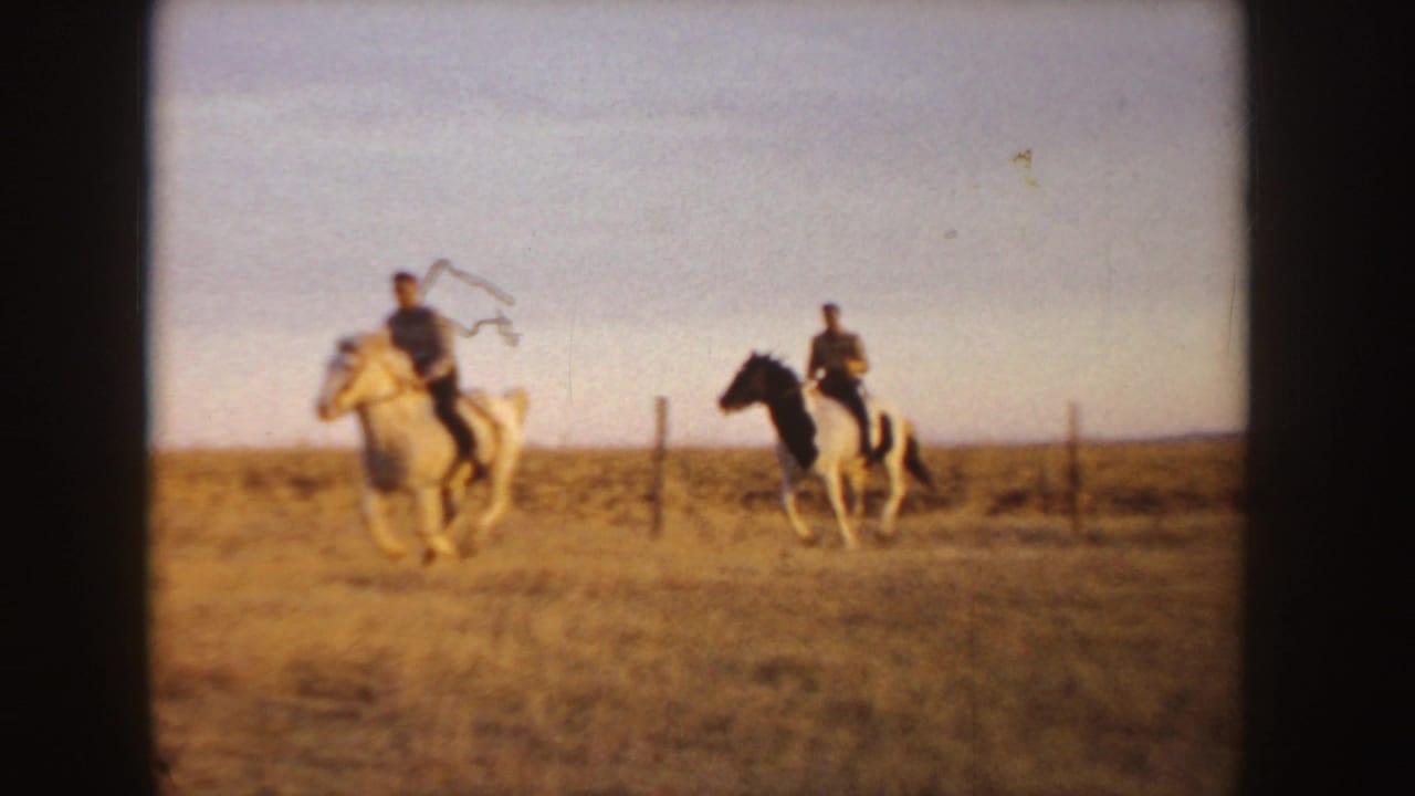 Two horseback riders cross a tan field near fence posts under open sky.