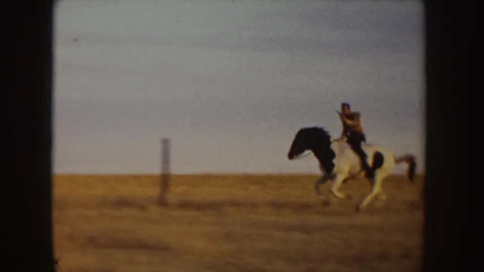 1955 Idaho 8mm frame with a rider on a black-and-white pinto crossing an open tan field beneath a pale sky.