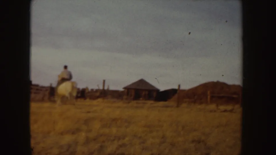 1955 Idaho 8mm frame showing a rider on a white horse near hay bales, fencing, and a small brown hut in a tan field.