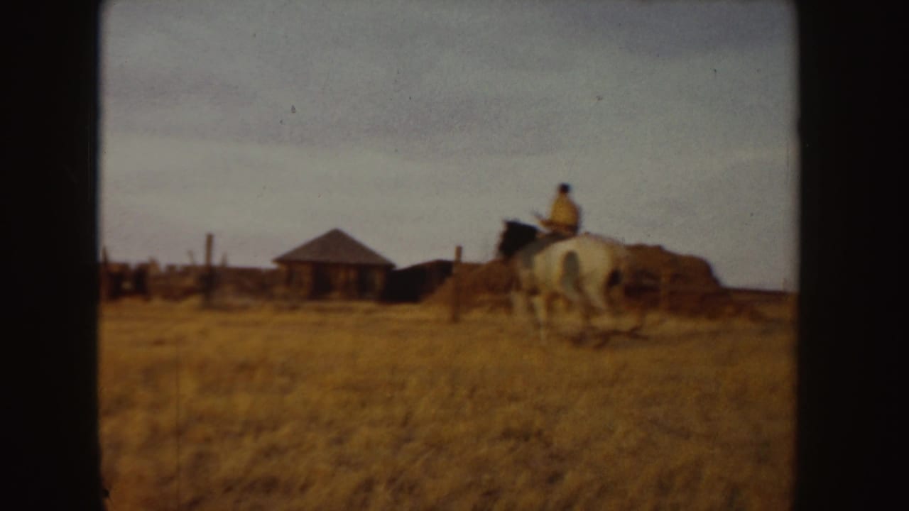1955 Idaho 8mm frame showing a rider on a white horse moving across dry pasture toward ranch buildings.