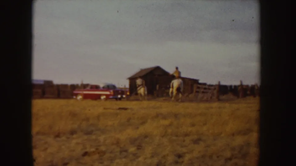 1955 Idaho home-movie frame with a horseback rider near a barn and a red parked car in the background.