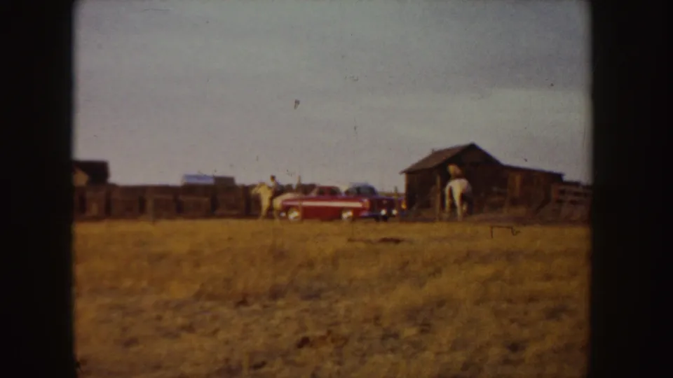 1955 Idaho ranch scene with distant riders, a red car, wooden barn, fences, and dry pasture.