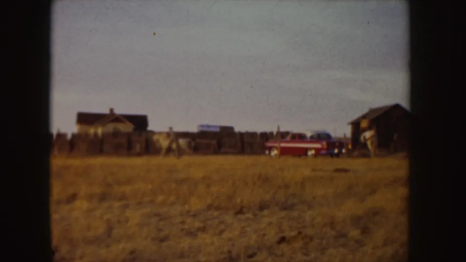 In 1955 Idaho home-movie footage, a distant rider on a white horse moves across a dry field near a wooden fence, outbuildings, and a red car.