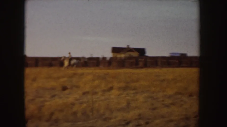 A distant person rides a white horse along a fenced rural field in Idaho, with modest buildings set back under a clear sky.