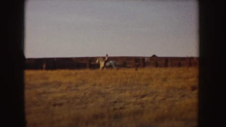The white horse and rider appear farther along the dry Idaho field, framed by a long fence and low rural buildings in soft 8mm color.