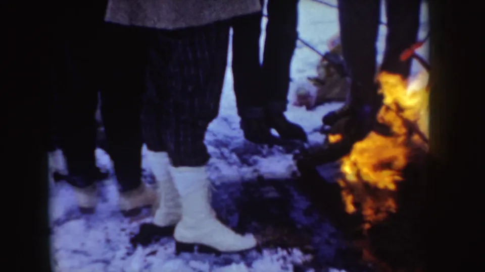 Bundled figures in snowy Idaho stand near a small outdoor fire, including a person in bright white winter boots.
