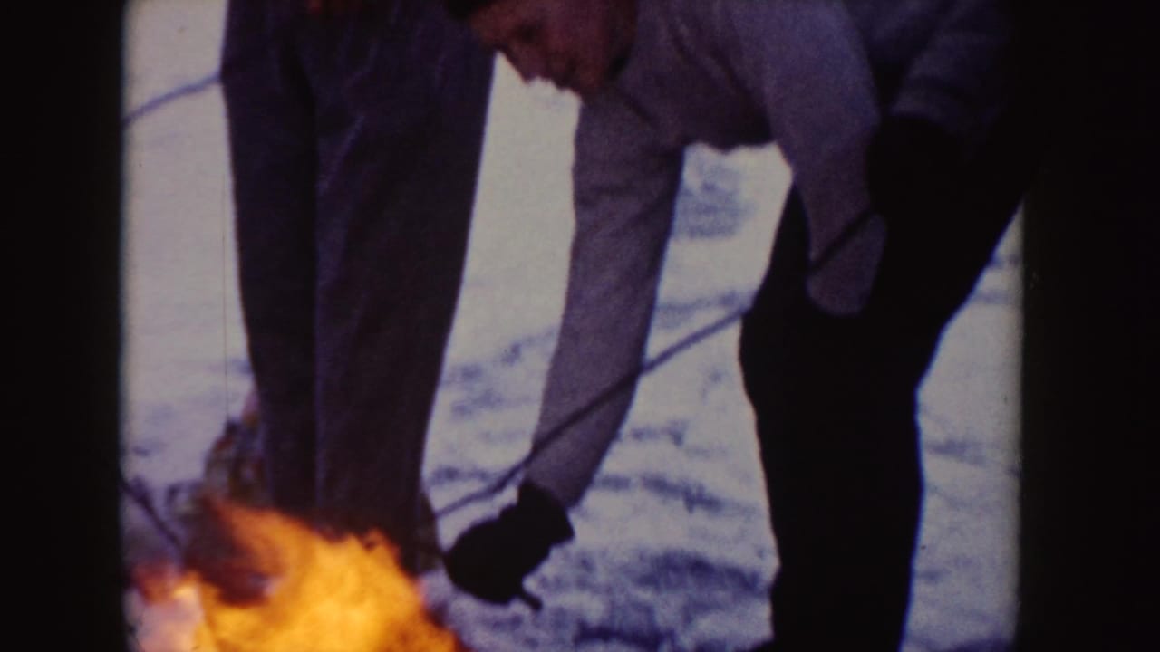 In 1955 Idaho home-movie footage, a young man in a gray sweater tends a small fire in snowy ground with roasting sticks nearby.