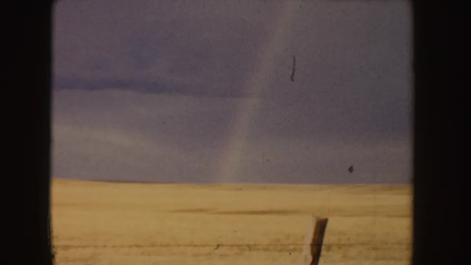 The 8mm frame shows a golden field, low horizon, cloudy sky, fence post, and faint rainbow in an Idaho landscape view.