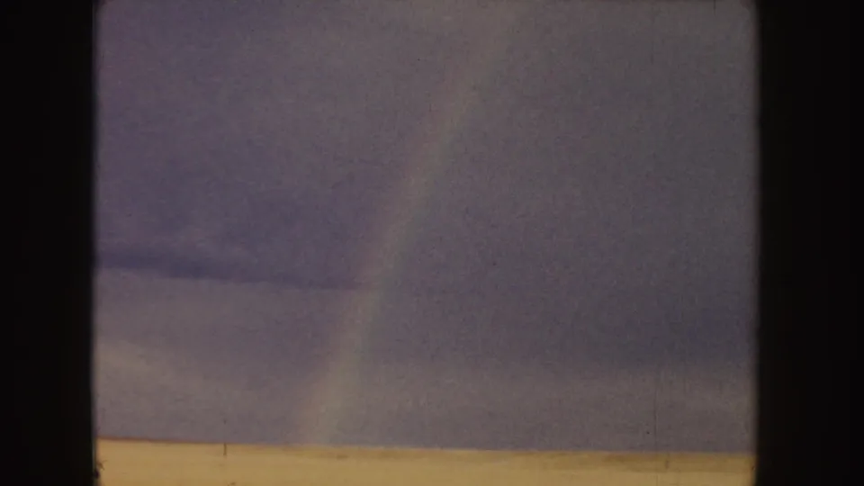 A soft, grainy landscape view shows cloudy sky above a pale field, with a faint rainbow band near the center-right.