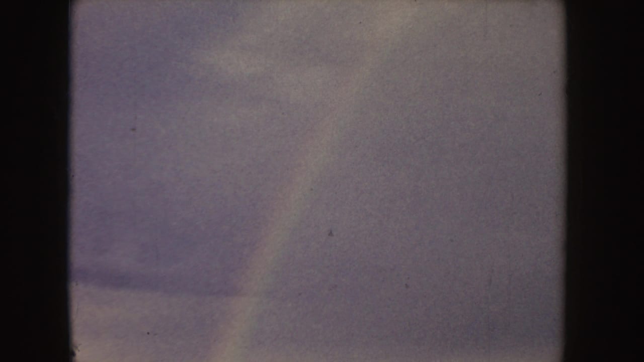 1955 Idaho 8mm frame showing a tan field beneath a blue-gray clouded sky, partly obscured by dark vertical frame edges.