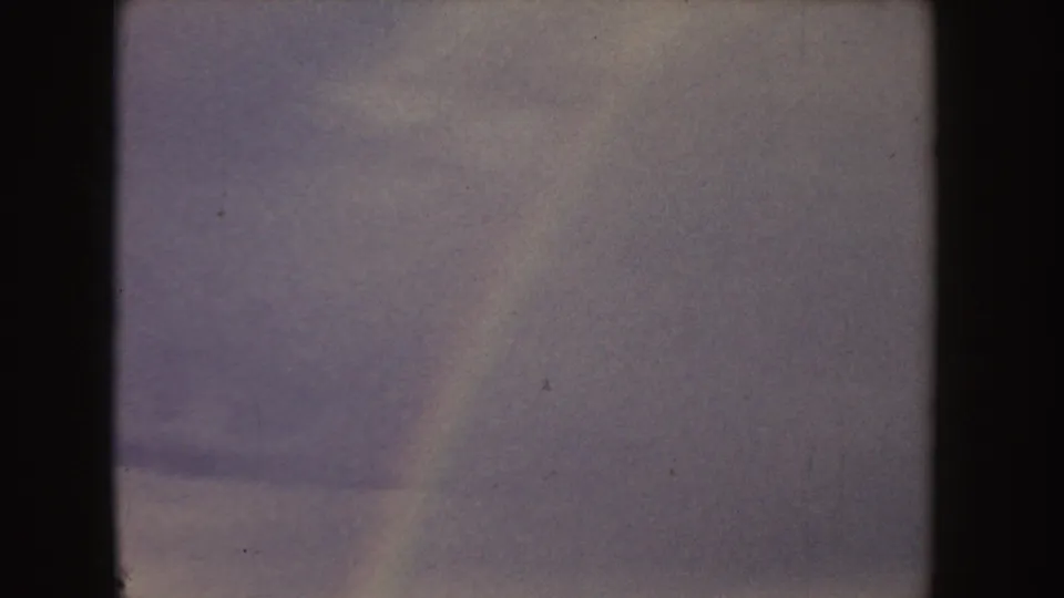 1955 Idaho 8mm frame showing a tan field beneath a blue-gray clouded sky, partly obscured by dark vertical frame edges.