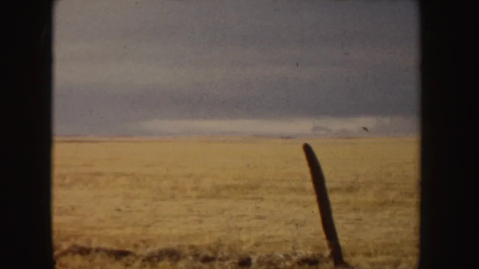 1955 Idaho home-movie frame with a leaning fence post in the foreground, open tan field, and heavy cloud cover beyond.