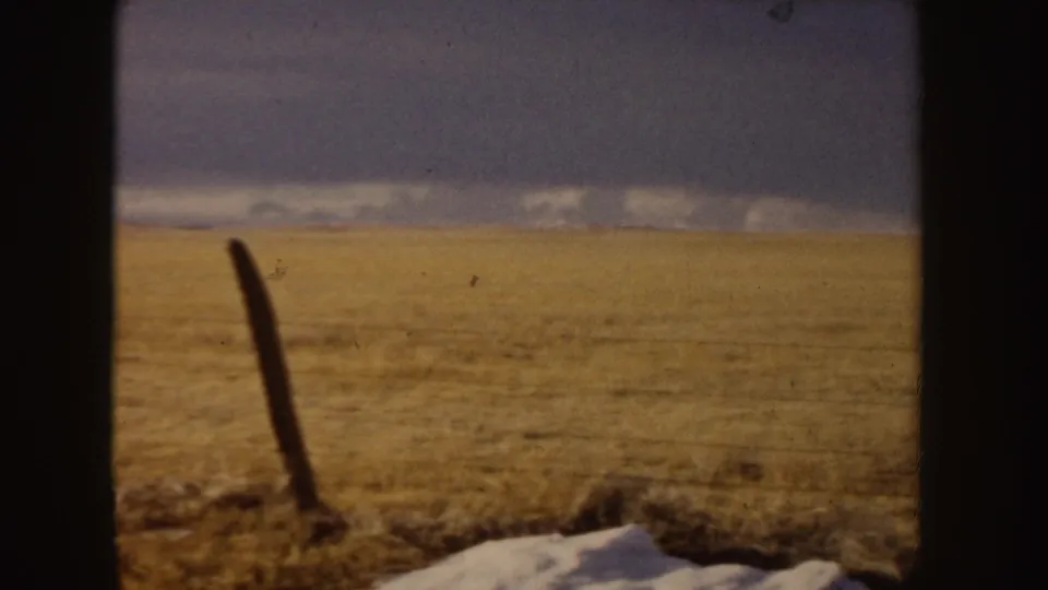 1955 Idaho 8mm frame showing a foreground fence post, pale ground or snow patch, tan field, and dark blue-gray clouds.