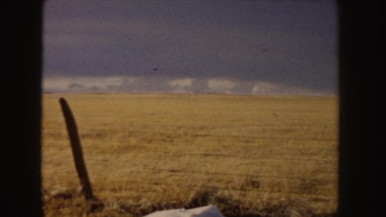 A 1955 8mm frame shows an open Idaho field or roadside view under a dark sky, partially blocked by vertical film or window edges.