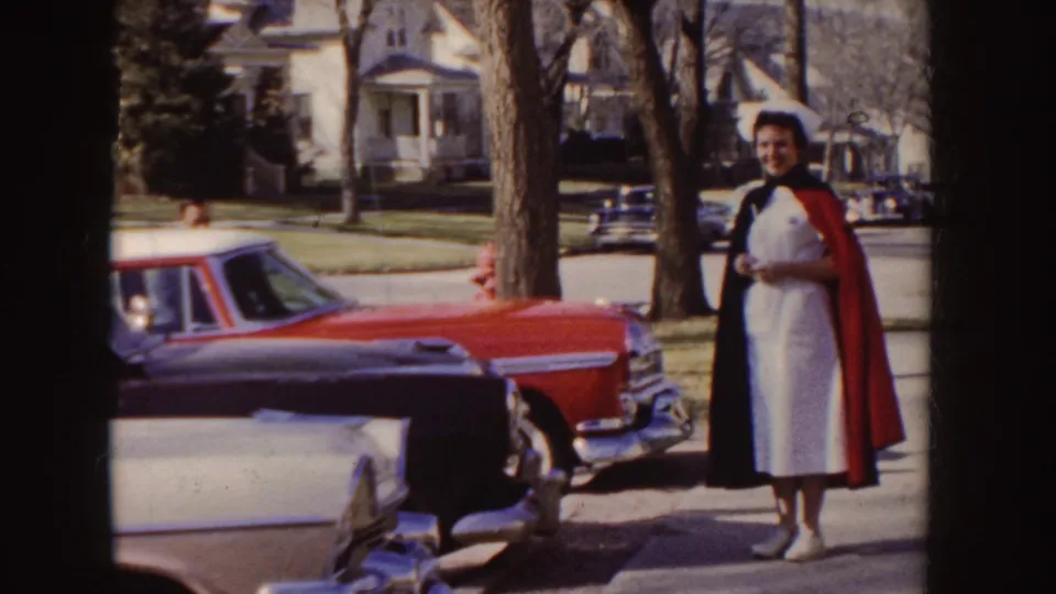 A nurse in a white uniform and dark cape stands near parked mid-century cars on a sunny Idaho residential street in 1955 footage.