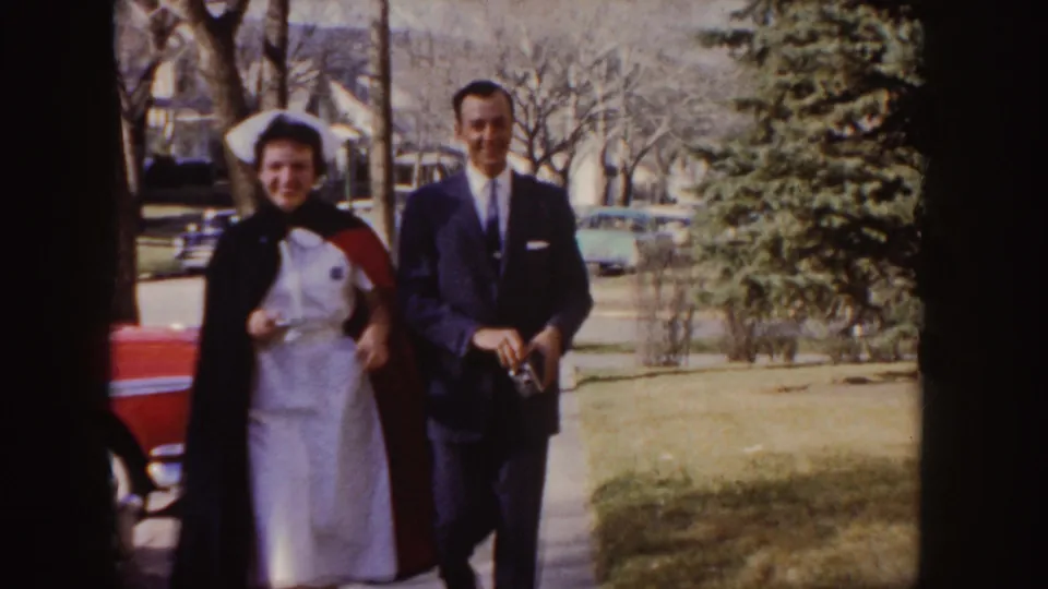 A 1955 Idaho home-movie frame shows a nurse in white with a dark cape walking beside a man in a grey suit on a suburban sidewalk.