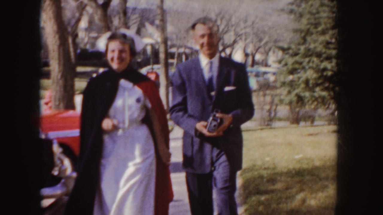 Young woman in nurse uniform and stole walks beside a suited man on a sidewalk.