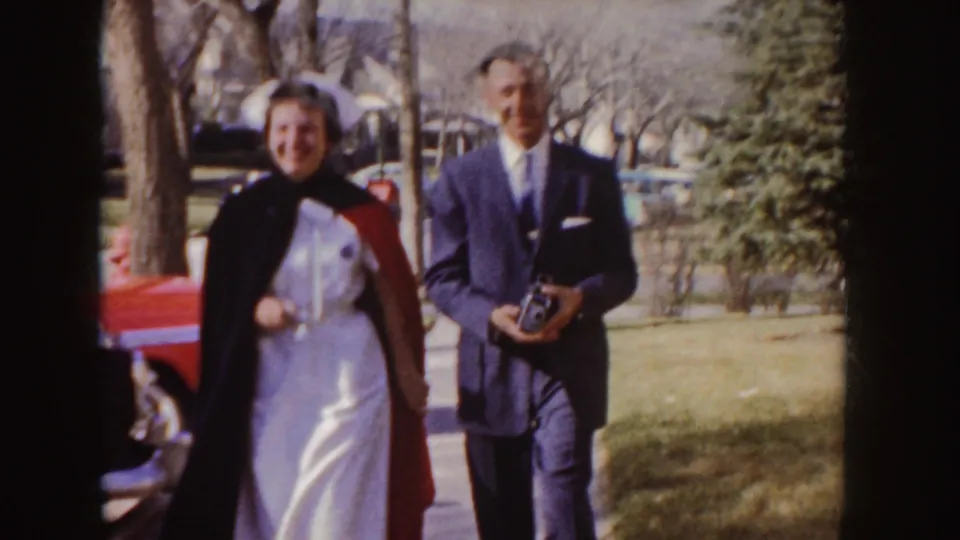 In 1955 Idaho home-movie footage, a young woman in a white nurse uniform and red-dark stole walks beside a man in a dark suit.