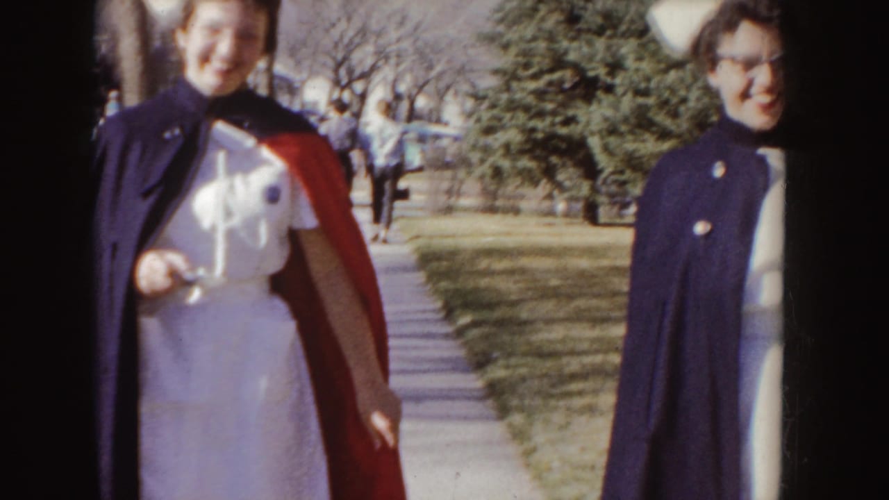 Smiling nurse graduate in a red and dark stole walks near another woman on a sidewalk.