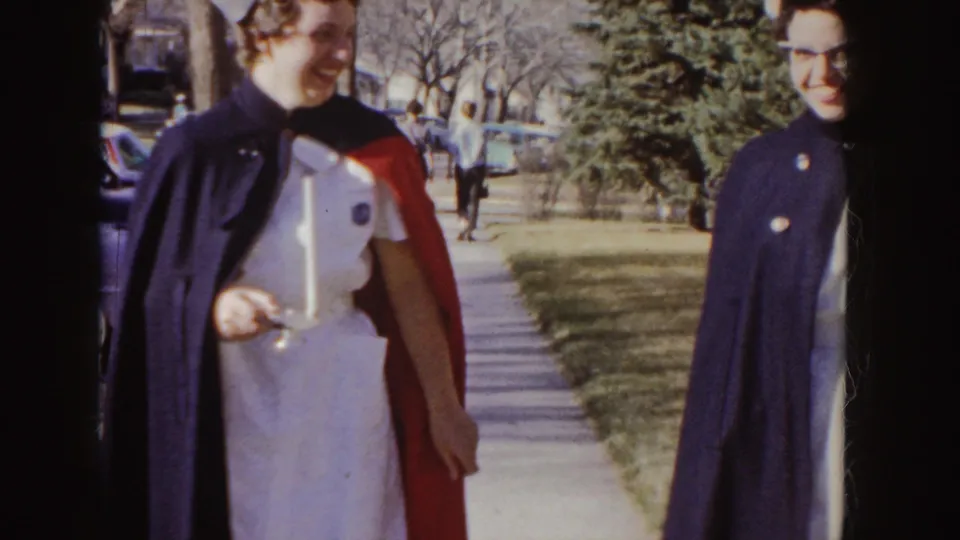 The nurse graduate in white and stole passes close to the 8mm camera with another woman in dark outerwear beside her.