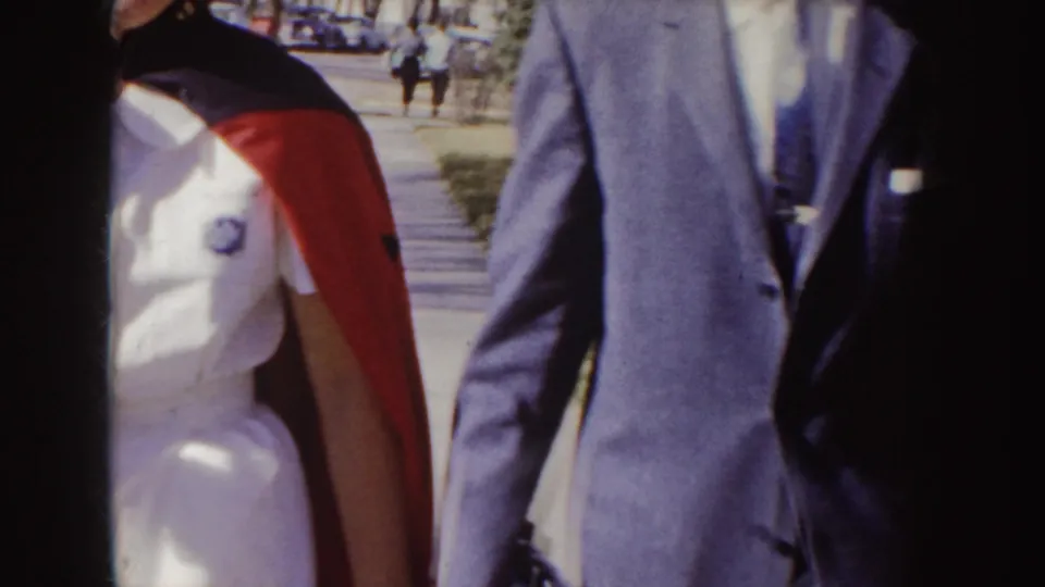 A close, partly cropped 8mm frame shows the nurse graduate's red-and-dark stole and the suited companion moving past the camera.