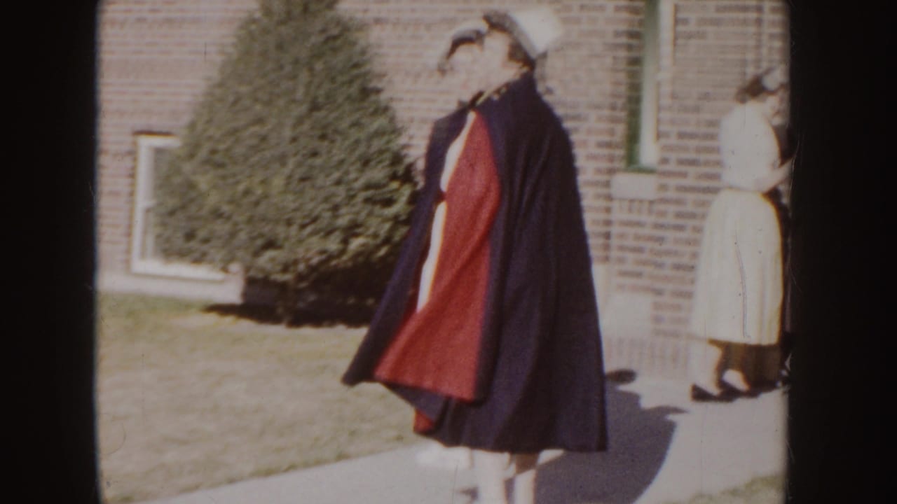 1955 Idaho home-movie frame of a nurse in a dark cape with red lining outside a brick building, with another person nearby.