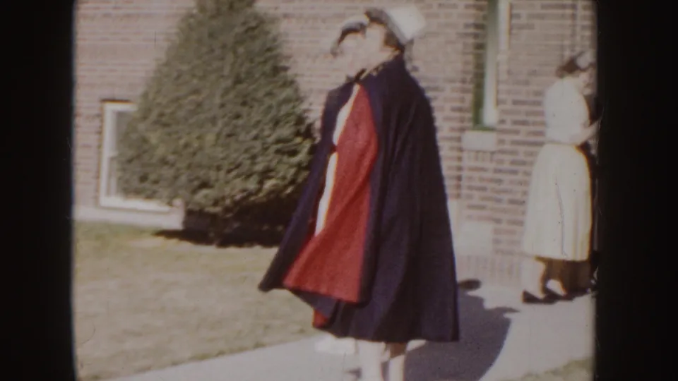 1955 Idaho home-movie frame of a nurse in a dark cape with red lining outside a brick building, with another person nearby.