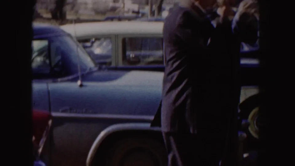 A parked mid-century car and partially visible person appear outside a brick building in 1955 Idaho footage.