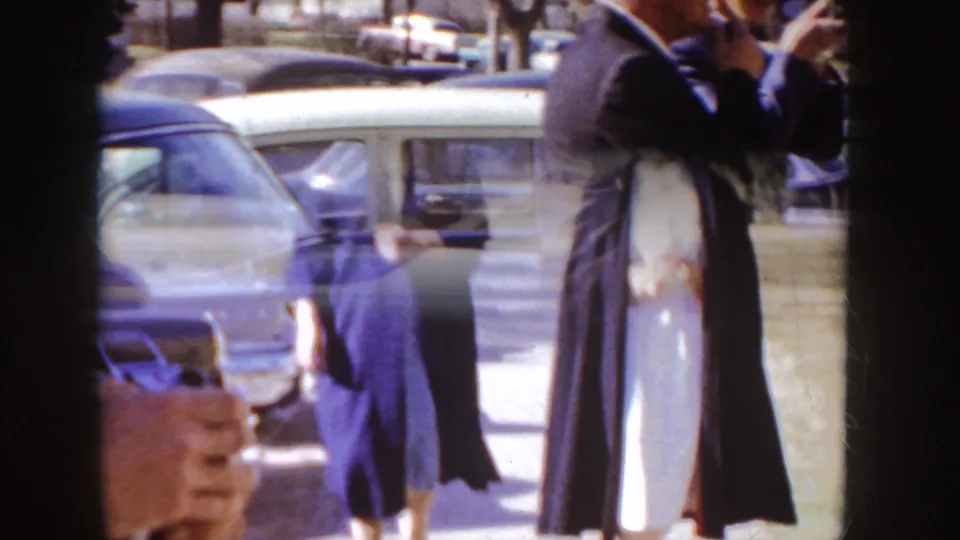 Two uniformed nurses in dark capes appear to embrace near parked mid-century cars outside a brick building in Idaho.