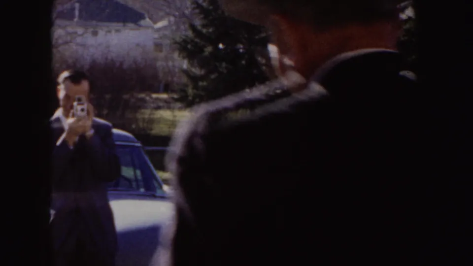 1955 Idaho frame with a man aiming a small camera outside, parked cars and houses behind him.